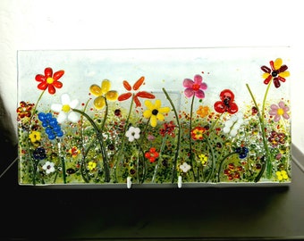 May include: A colorful glass art piece depicting a field of flowers. The flowers are made of glass and are in various colors, including red, yellow, blue, pink, and white. The background is a light blue.