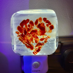 Puede incluir: Una luz nocturna blanca con un inserto de vidrio transparente en forma de corazón. El corazón está lleno de un patrón de remolino de rojo y naranja.