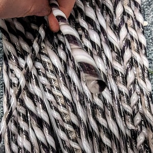 Puede incluir: Primer plano de un conjunto de varillas de vidrio retorcidas. Las varillas son principalmente blancas con rayas negras y moradas, creando un patrón a rayas. Una mano sostiene una de las varillas, mostrando el diseño retorcido. Las varillas parecen ser parte de un proyecto de artesanía.