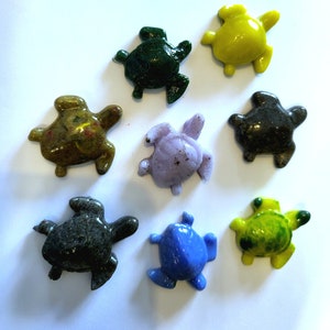 May include: Eight small, turtle-shaped figurines in various colours. The figurines include shades of green, yellow, blue, purple, and brown. Each turtle has a smooth, glossy finish and is arranged on a white surface.