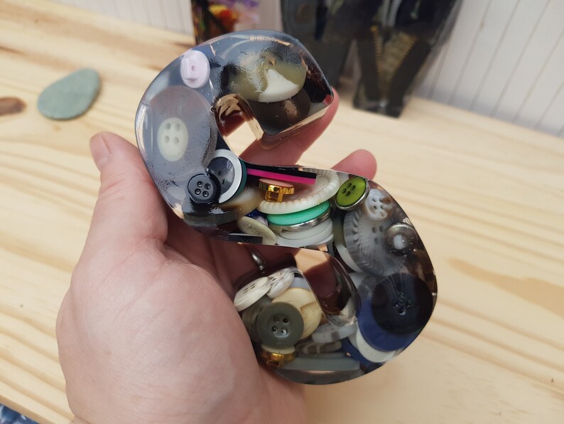 May include: A clear resin letter 'S' filled with a variety of colorful buttons. The buttons are arranged in a random pattern and are visible through the resin.