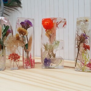 May include: The word "MEL" spelled out in clear resin letters, each containing a variety of dried flowers and foliage. The letters are arranged on a light wood surface.
