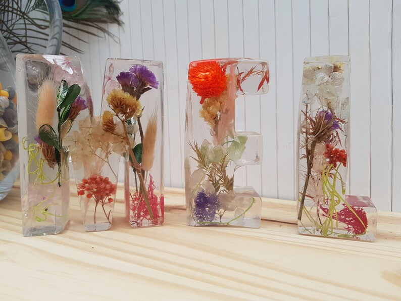 May include: The word "MEL" spelled out in clear resin letters, each containing a variety of dried flowers and foliage. The letters are arranged on a light wood surface.