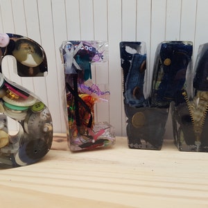 May include: Resin letters spelling "SEW" with various sewing-themed items embedded inside. The "S" is filled with buttons, the "E" with colorful ribbons and thread, and the "W" with denim fabric and a zipper.