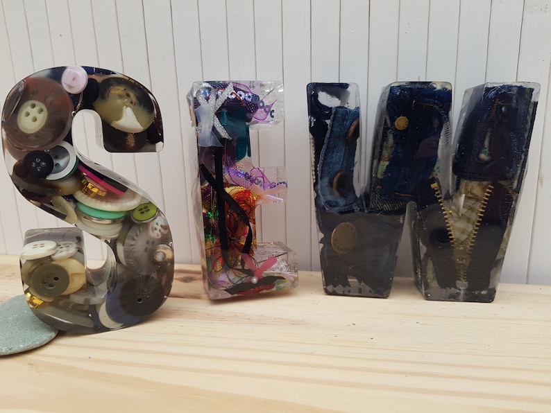 May include: Resin letters spelling "SEW" with various sewing-themed items embedded inside. The "S" is filled with buttons, the "E" with colorful ribbons and thread, and the "W" with denim fabric and a zipper.