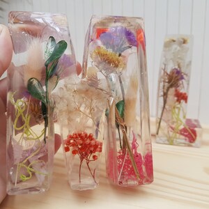 May include: Three clear resin letters, M, A, and M, each filled with dried flowers and greenery. The letters are arranged in a row on a light wood surface.