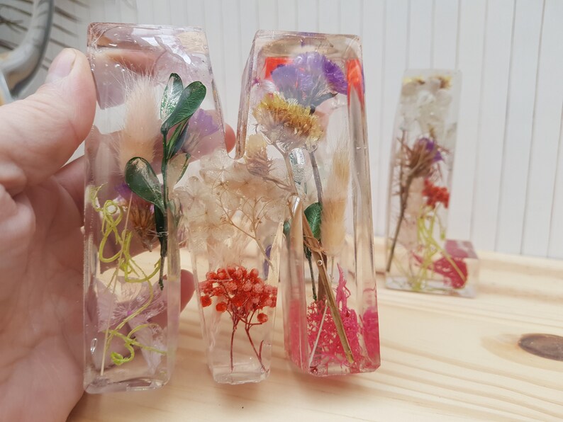 May include: Three clear resin letters, M, A, and M, each filled with dried flowers and greenery. The letters are arranged in a row on a light wood surface.
