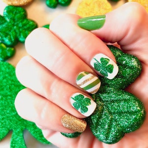 May include: Close-up of a hand with green and gold nail polish. The nails are decorated with shamrock designs and glitter.