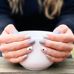 May include: A close-up of a person's hands holding a white ceramic mug. The person's fingernails are painted with a light gray nail polish and have white and black heart designs.