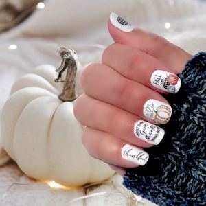May include: White nail polish with fall-themed designs. The designs include pumpkins, leaves, and the words "Fall", "Give Thanks", "Hello Fall", and "Thankful".