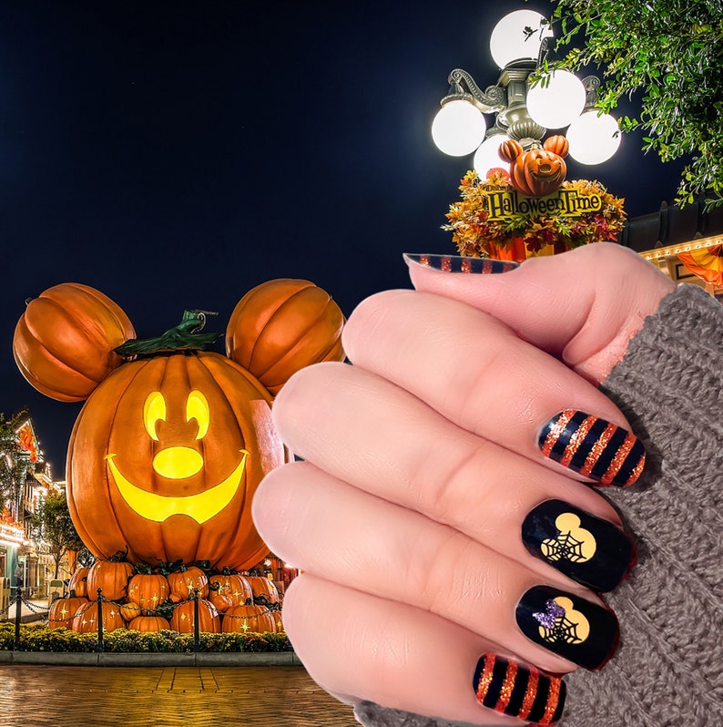 May include: A hand with black and orange Halloween themed nail art featuring Mickey Mouse silhouettes. The nail art is displayed in front of a large jack-o-lantern with Mickey Mouse ears.
