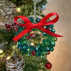以下が含まれることがあります： 緑と赤のビーズで作られたリース型のクリスマスツリーオーナメント。鮮やかな赤いリボンで飾られています。リースは銀色のチェーンで吊り下げられており、背景には他のオーナメントと木の枝があります。