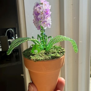 Pode incluir: Uma flor de jacinto de contas artesanal num vaso de terracota. A flor apresenta contas roxas e transparentes, folhas de contas verdes e uma pequena figura de rã verde. O vaso é preenchido com musgo verde.