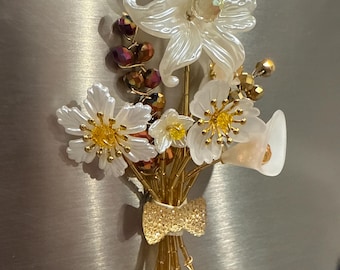 May include: A decorative bouquet of white and yellow flowers with gold accents and a gold bow. The bouquet includes lilies, calla lilies, and other floral elements, all tied together with gold wire.