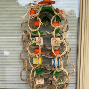 May include: A bird toy with a variety of textures and colors. It features wooden blocks with letters and images, orange spheres, green and brown leaf-shaped elements, and woven vine-like structures. The toy is suspended from a metal chain.