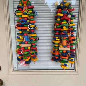 May include: Two vertical hanging bird toys constructed from a variety of colorful wooden shapes, including blocks, cylinders, and spheres, interspersed with plastic rings and beads, all strung together on a sturdy cord for suspension.