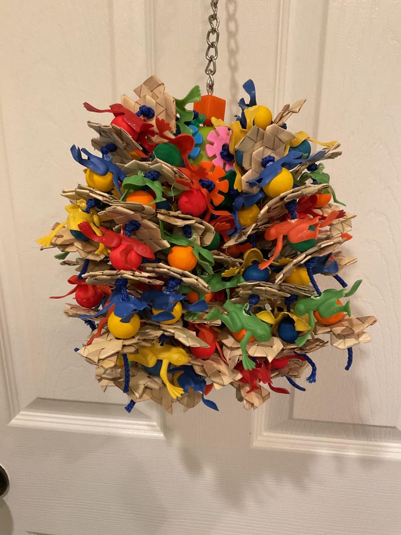May include: A colorful bird toy for parrots and other birds. The toy is made of natural woven material and features a variety of plastic toys, including balls, rings, and figures. The toy is attached to a metal chain for hanging.