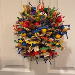 May include: A colorful bird toy made of natural materials and plastic. The toy is a sphere with a variety of textures and colors, including blue, red, yellow, green, and brown. The toy is hanging from a chain.
