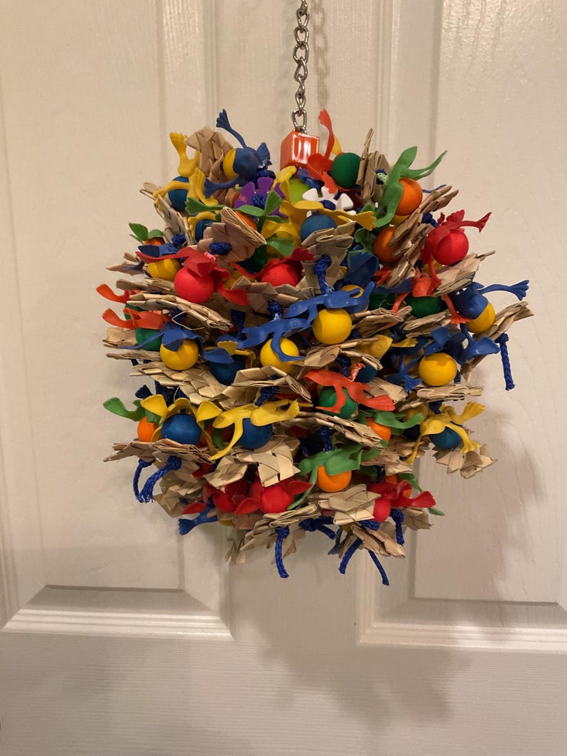 May include: A colorful bird toy made of natural materials and plastic. The toy is a sphere with a variety of textures and colors, including blue, red, yellow, green, and brown. The toy is hanging from a chain.