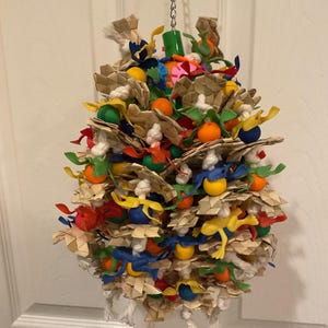 May include: A colorful bird toy made of woven palm leaves, rope, and plastic beads. The toy is filled with various shapes and colors, including green, blue, yellow, orange, and red. It is hanging from a silver chain.