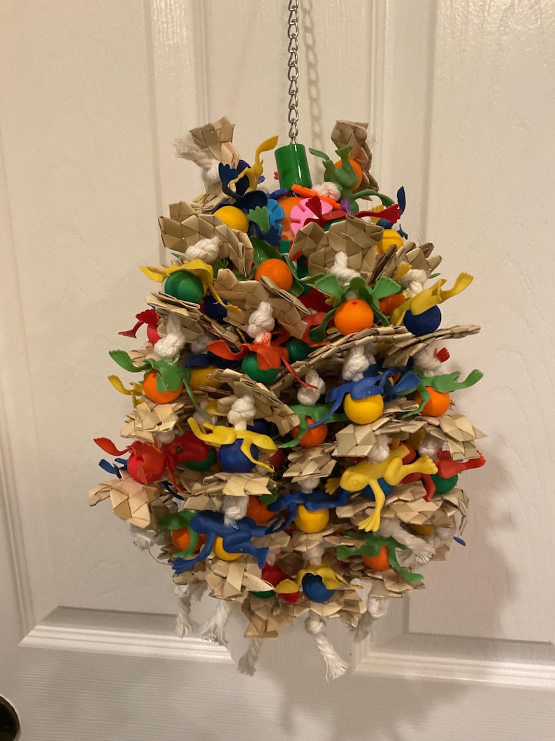 May include: A colorful bird toy made of woven palm leaves, rope, and plastic beads. The toy is filled with various shapes and colors, including green, blue, yellow, orange, and red. It is hanging from a silver chain.