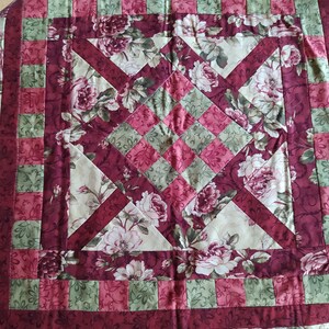 Könnte beinhalten: Ein gesteppter Tischläufer mit einem floralen Muster in Grün-, Rosa- und Burgunder-Tönen. Der Läufer hat ein Rautenmuster mit einem zentralen Quadrat.