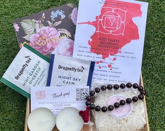 May include: A gift box filled with self-care items, including tea bags, bath salts, candles, a bracelet, and a card with affirmations. The box is on a green grassy surface.