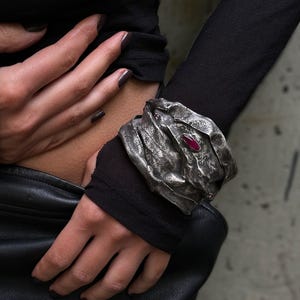 May include: A wide, textured silver cuff bracelet with a dark, oxidised finish and a single, elongated, deep red gemstone set into the centre. The metal surface features a crinkled, organic appearance with irregular folds and ridges throughout.