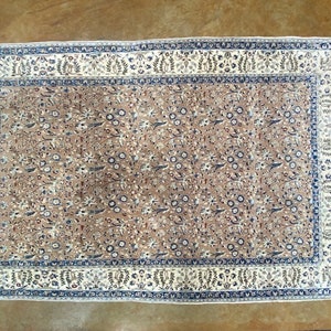 May include: A beige rug with a floral pattern in shades of blue, brown, and white. The rug has a border with a repeating pattern of leaves and flowers.