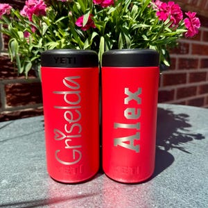 May include: Two red insulated drink holders with black lids. One holder has the name "Griselda" in silver script with a heart, and the other has the name "Alex" in silver block letters. The brand name "YETI" is at the bottom of each holder.