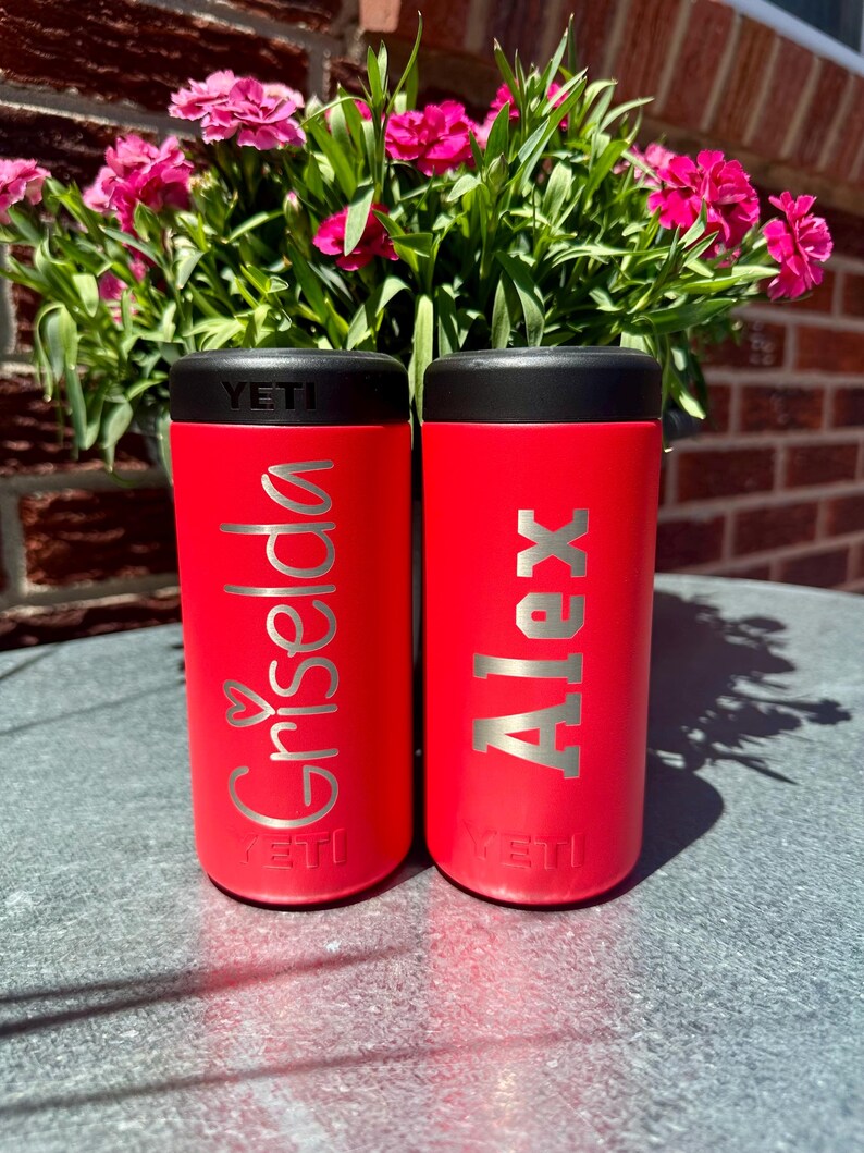 May include: Two red insulated drink holders with black lids. One holder has the name "Griselda" in silver script with a heart, and the other has the name "Alex" in silver block letters. The brand name "YETI" is at the bottom of each holder.