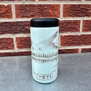 May include: A light blue YETI can cooler with a black top. The cooler has a mountain and lake scene etched in silver. The YETI logo is at the bottom. The background is a red brick wall.