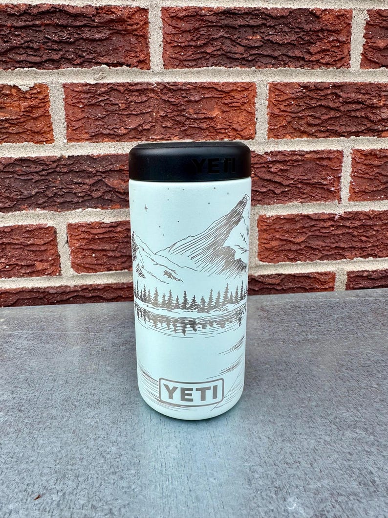 May include: A light blue YETI can cooler with a black top. The cooler has a mountain and lake scene etched in silver. The YETI logo is at the bottom. The background is a red brick wall.