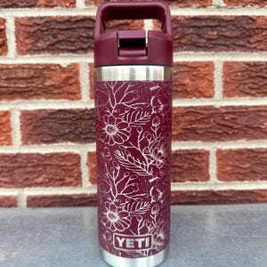 May include: A maroon YETI water bottle featuring a white line-art floral pattern with various flowers, leaves, and butterflies. The bottle has a metallic silver rim at the top and bottom, and a maroon plastic lid with an integrated carrying handle.