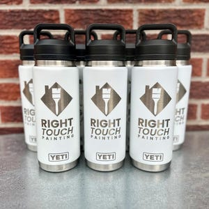 Puede incluir: Un grupo de botellas de agua aisladas blancas con tapas y asas negras. Cada botella presenta el texto "RIGHT TOUCH PAINTING" y un logotipo de una casa y un pincel. También es visible la marca "YETI".