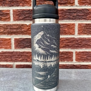 May include: A dark grey YETI water bottle with a black handle lid. The bottle features a laser-engraved mountain landscape design with a forest, a lake, and a single star above the peak. The surface has a textured, speckled finish.