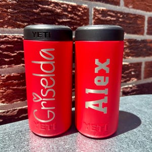 May include: Two red insulated tumblers with black lids. One tumbler has the name "Griselda" in silver script with a heart, and the other has the name "Alex" in block letters. Both tumblers have the "YETI" logo.