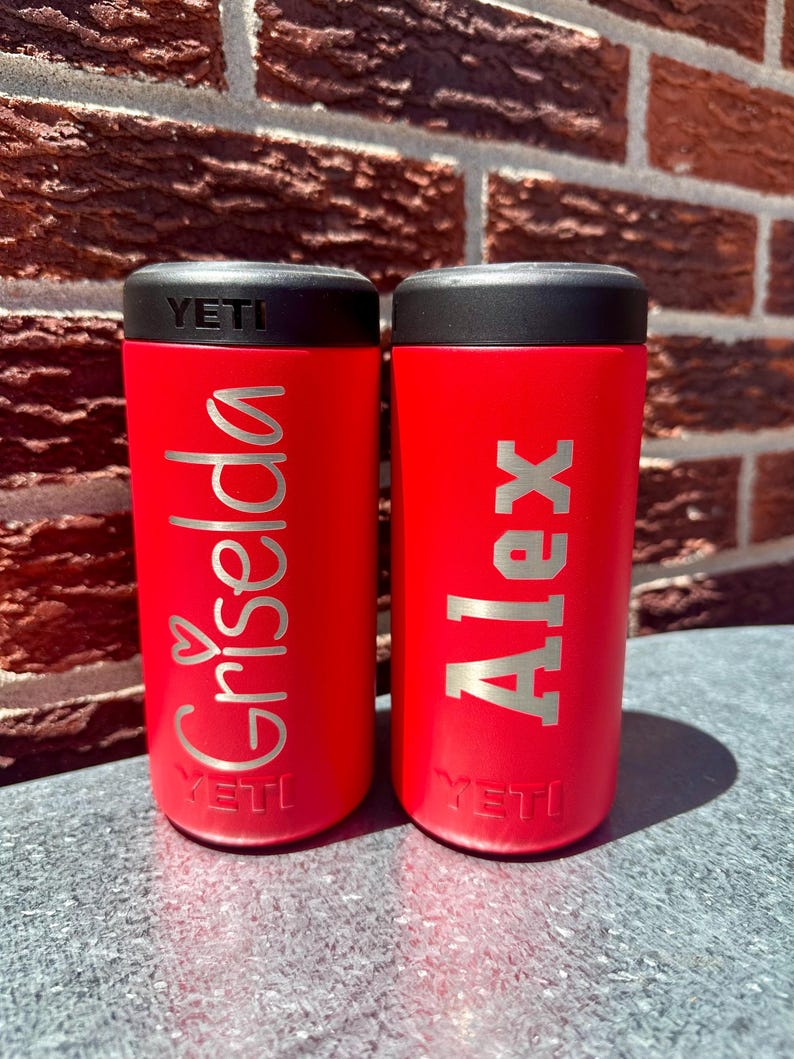 May include: Two red insulated tumblers with black lids. One tumbler has the name "Griselda" in silver script with a heart, and the other has the name "Alex" in block letters. Both tumblers have the "YETI" logo.