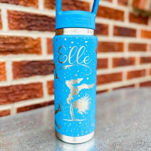 Pode incluir: Uma garrafa de água YETI azul brilhante com um design gravado em prata apresentando o nome Elle em letra cursiva, rodeado por estrelas prateadas e a silhueta de uma ginasta a fazer pino. A garrafa tem uma tampa azul com pega.