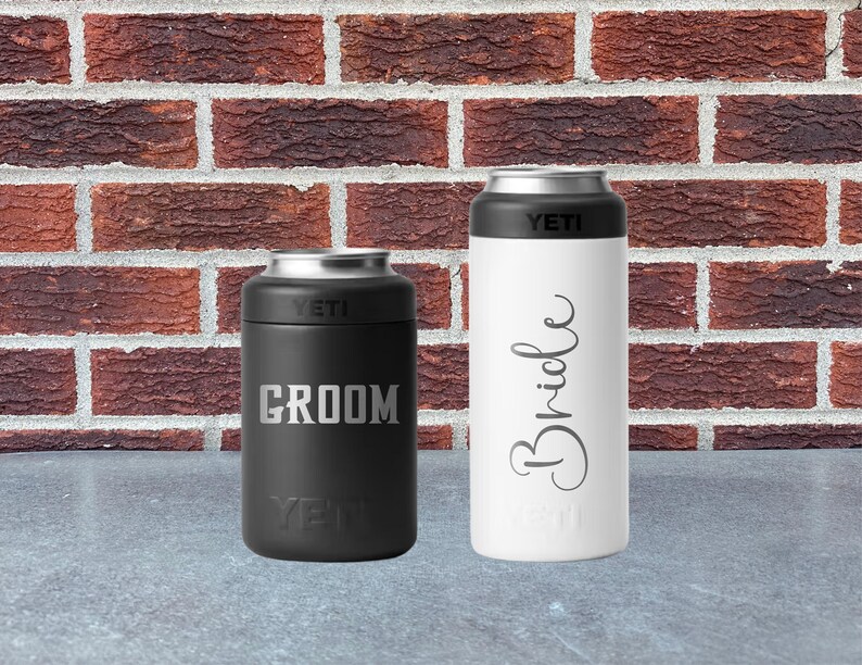 May include: Two insulated beverage holders, one black and one white, with silver rims. The black holder has "Groom" in silver lettering, and the white holder has "Bride" in cursive. Both have the "YETI" logo.