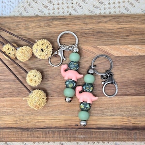 May include: Two keychains with pink dinosaur charms and green and patterned beads. The keychains have silver-colored hardware, including a clasp and a keyring. The keychains are displayed on a wooden surface with decorative dried flowers.