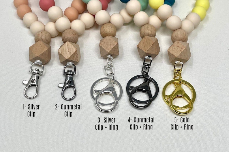 Puede incluir: Cinco llaveros diferentes con cuentas de madera y clips y anillos de metal. Los llaveros est&aacute;n etiquetados como 1- Clip plateado, 2- Clip de metal, 3- Clip plateado + Anillo, 4- Clip de metal + Anillo y 5- Clip dorado + Anillo.