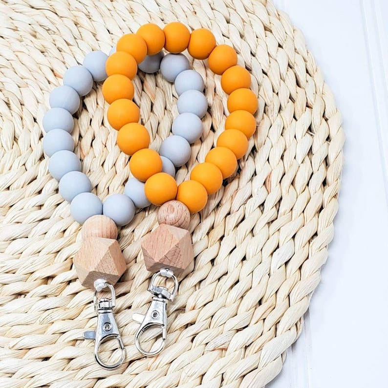 Puede incluir: Dos llaveros de cuentas de silicona, uno gris y otro naranja, con cuentas de madera y clips plateados.