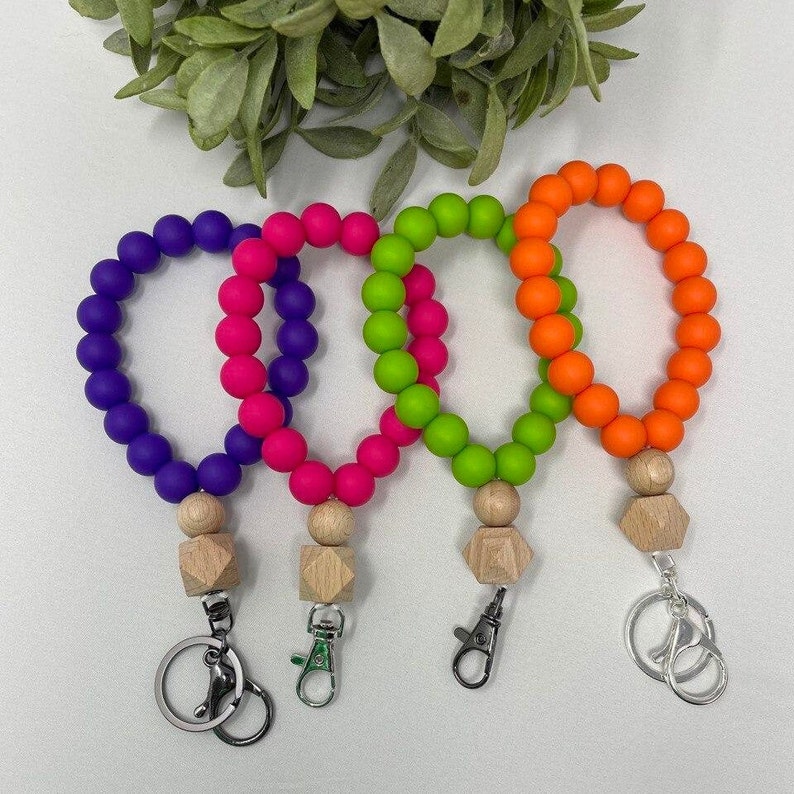 Puede incluir: Cuatro llaveros de cuentas de silicona con dijes de madera y clips met&aacute;licos. Los llaveros son de color morado, rosa, verde y naranja.