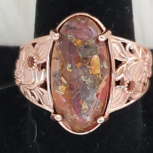 May include: Rose gold ring with an oval-shaped, multi-colored stone. The stone features shades of red, brown, and gold. The ring band has a floral design with intricate cut-out details. The ring is displayed on a black surface.