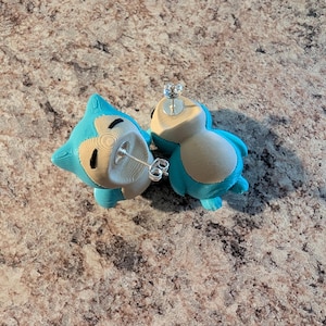 Pode incluir: Um par de brincos de tarracha com o formato da personagem Snorlax, apresentando um esquema de cores azul e bege com um acabamento texturizado. Cada brinco inclui um pino de metal prateado e uma tarracha borboleta para fixação segura no lóbulo.