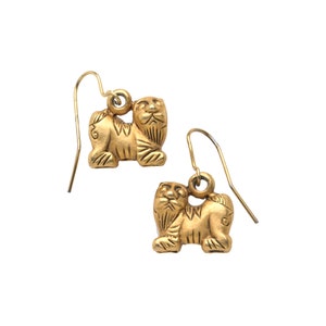 May include: A pair of gold-tone earrings featuring a detailed lion design. The lion is facing forward with its head tilted slightly to the side. The earrings are suspended from a simple wire loop.