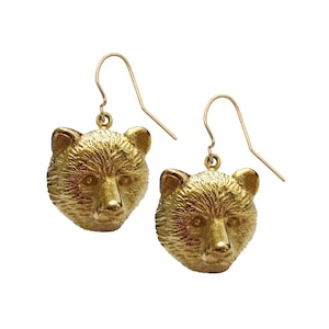 May include: A pair of gold-colored bear head earrings. The earrings feature detailed bear faces with textured fur and are suspended from gold-colored ear wires. The bear heads are facing forward, showcasing their features.