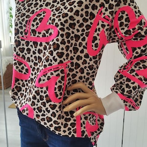 Könnte beinhalten: Ein langärmliges Oberteil mit Leopardenmuster und dem Wort "LOVE" in leuchtendem Pink. Das Oberteil hat weiße Manschetten und einen weißen Ausschnitt. Das Model trägt eine blaue Jeans.