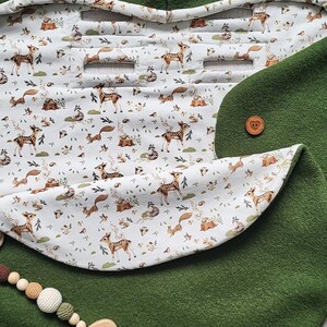 May include: A green baby stroller blanket with a hood and a woodland animal print lining. The blanket features a button closure and a wooden elephant-shaped teether with a beaded string. The lining displays deer, foxes, and other forest creatures.